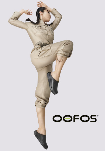 Athletic woman mid-air in tan jumpsuit and black clogs against grey backdrop, showcasing dynamic pose and footwear brand OOFOS logo.