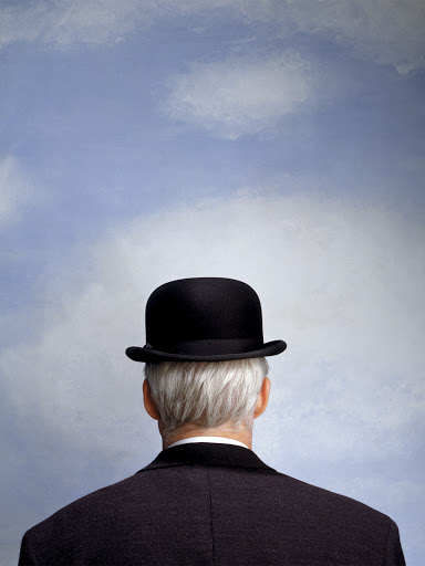Back of an older man with gray hair wearing a black bowler hat and suit against a blue sky with clouds in an eye-level shot.