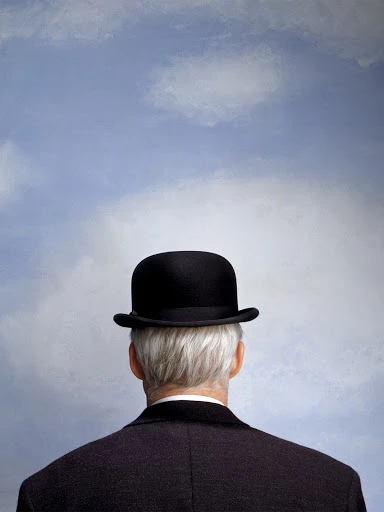Back of an older man with gray hair wearing a black bowler hat and suit against a blue sky with clouds in an eye-level shot.