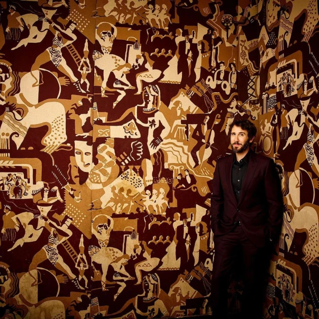 Man in a suit standing against a wall featuring a large art deco style illustration with various figures and geometric patterns in brown and beige tones.