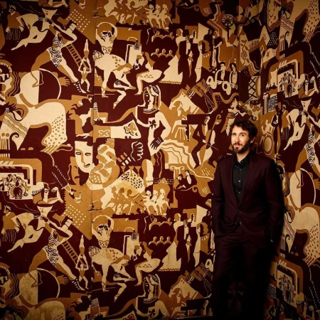 Man in a suit standing against a wall featuring a large art deco style illustration with various figures and geometric patterns in brown and beige tones.