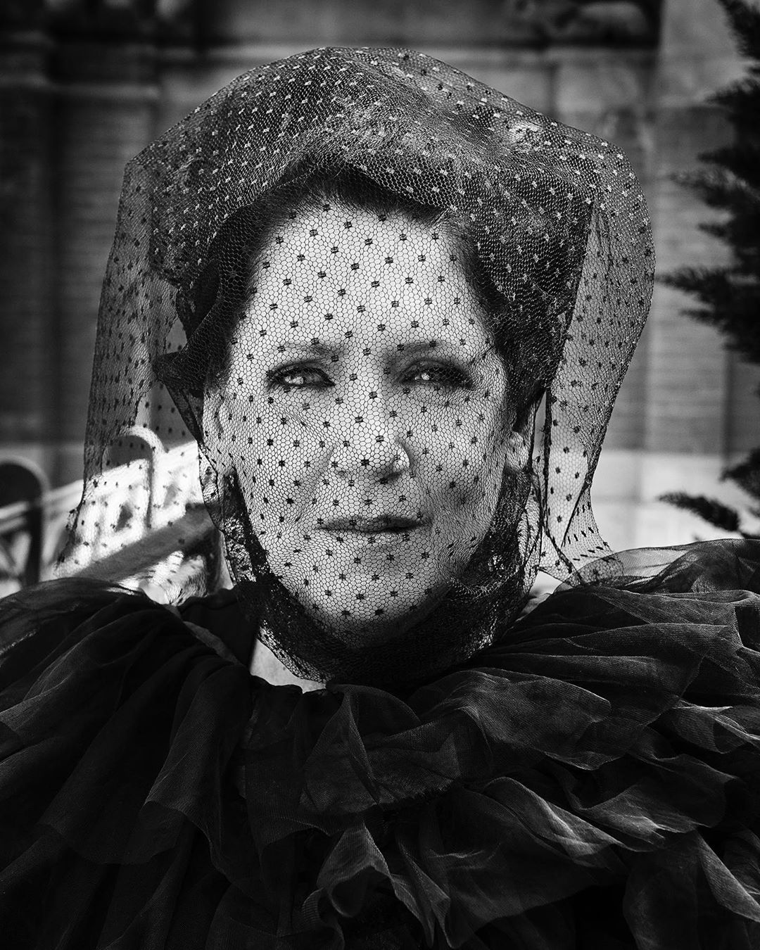 Close-up black and white portrait of an adult female wearing a black polka-dot veil and black ruffled collar with architectural background.