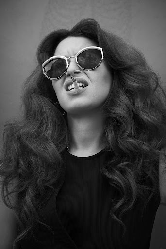 Black and white portrait of a stylish woman with curly hair wearing sunglasses and holding a cigarette in her mouth.