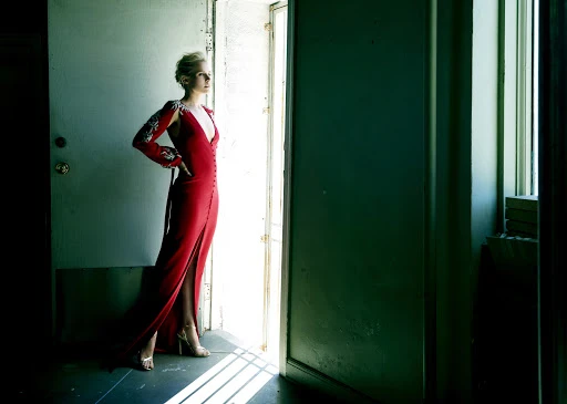 Woman in red gown poses in doorway with silver heels, arm raised, looking out into bright light. Indoor fashion photography.