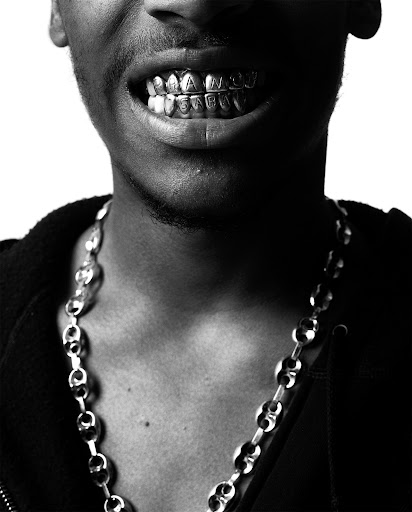 Close-up of a black man's mouth with custom gold teeth grills, reading "HANG US SLOW," adorned with a silver chain necklace.