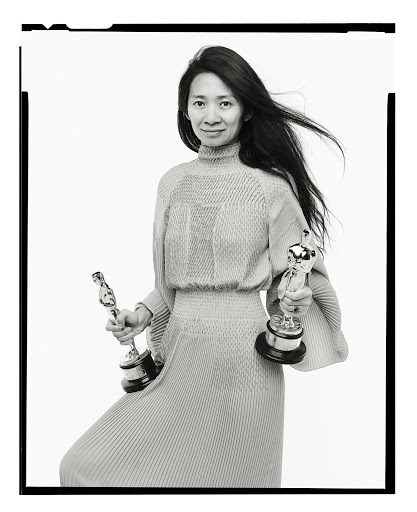 Black and white portrait of woman holding two awards. Long hair, high-neck dress, smiling in front of white background. Classic, elegant.