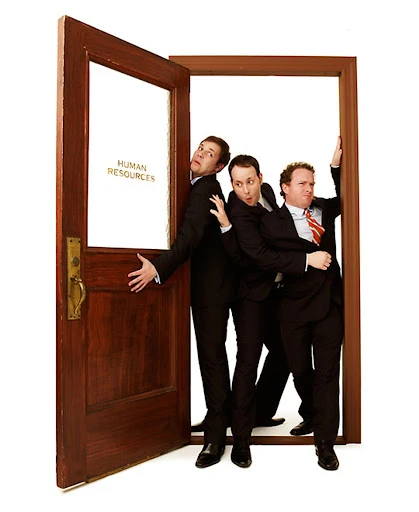 Three adult males crammed in a wooden door frame labeled “Human Resources” against a white backdrop, wearing dark suits.