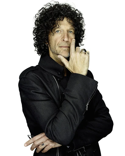 Portrait of an adult male with dark curly hair, wearing a black jacket, his fingers touching his face, against a white backdrop.