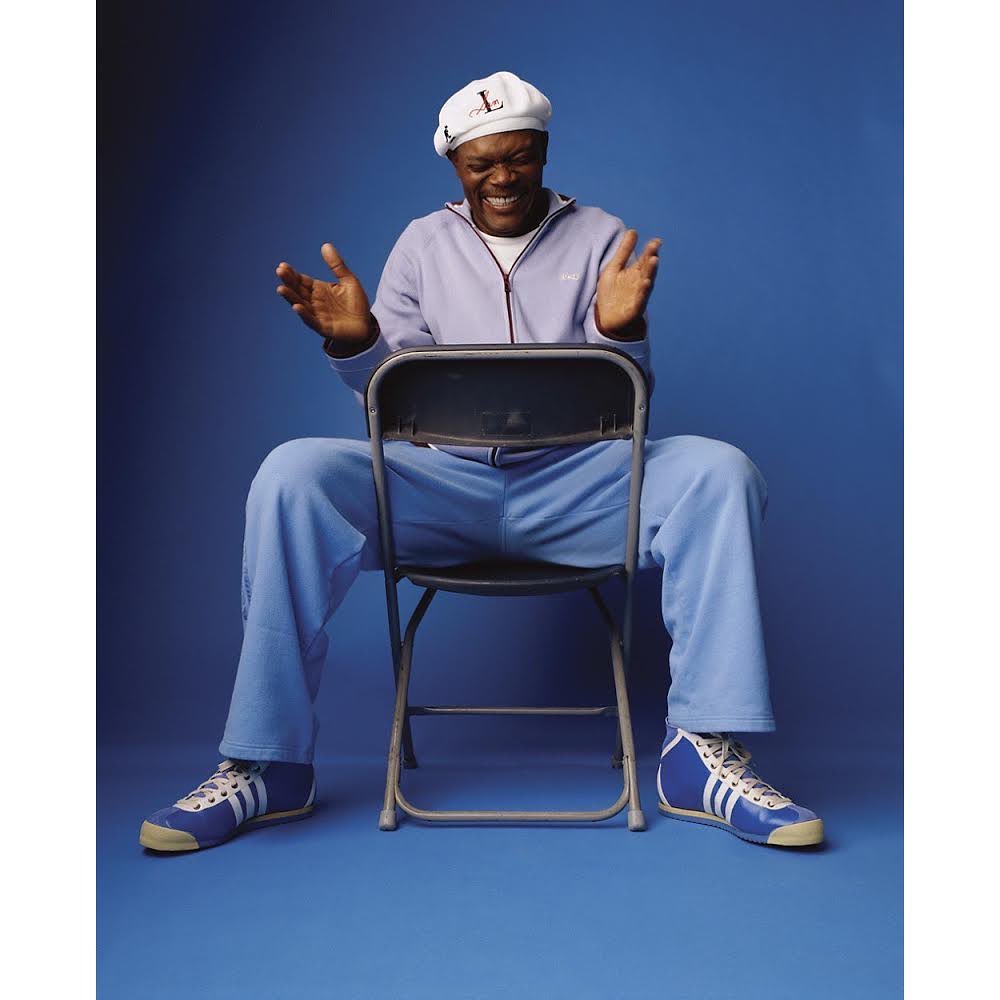 Adult male sitting on a chair in a studio, wearing a hat, light blue jogging suit, and blue-and-white tennis shoes.