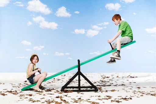 Two children, a girl with a crown and a red-haired boy, are playing on a green seesaw against a blue cloudy sky backdrop.