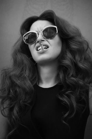 Monochrome portrait of a woman with long dark wavy hair wearing cat-eye sunglasses and holding a cigarette in her mouth