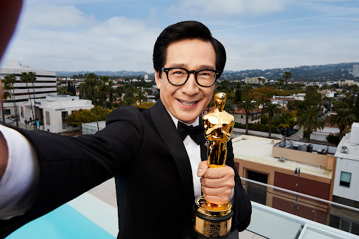 Smiling man in tuxedo holds a golden Oscar statue on rooftop with cityscape, palm trees, and buildings in the background.
