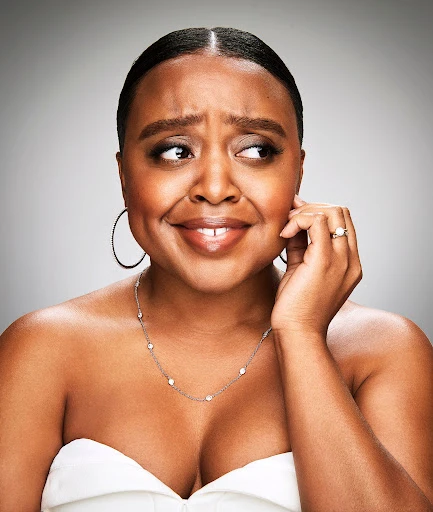 Headshot of a smiling dark-skinned woman with pulled-back hair, hoop earrings, and a silver necklace, posing in front of a grey background.