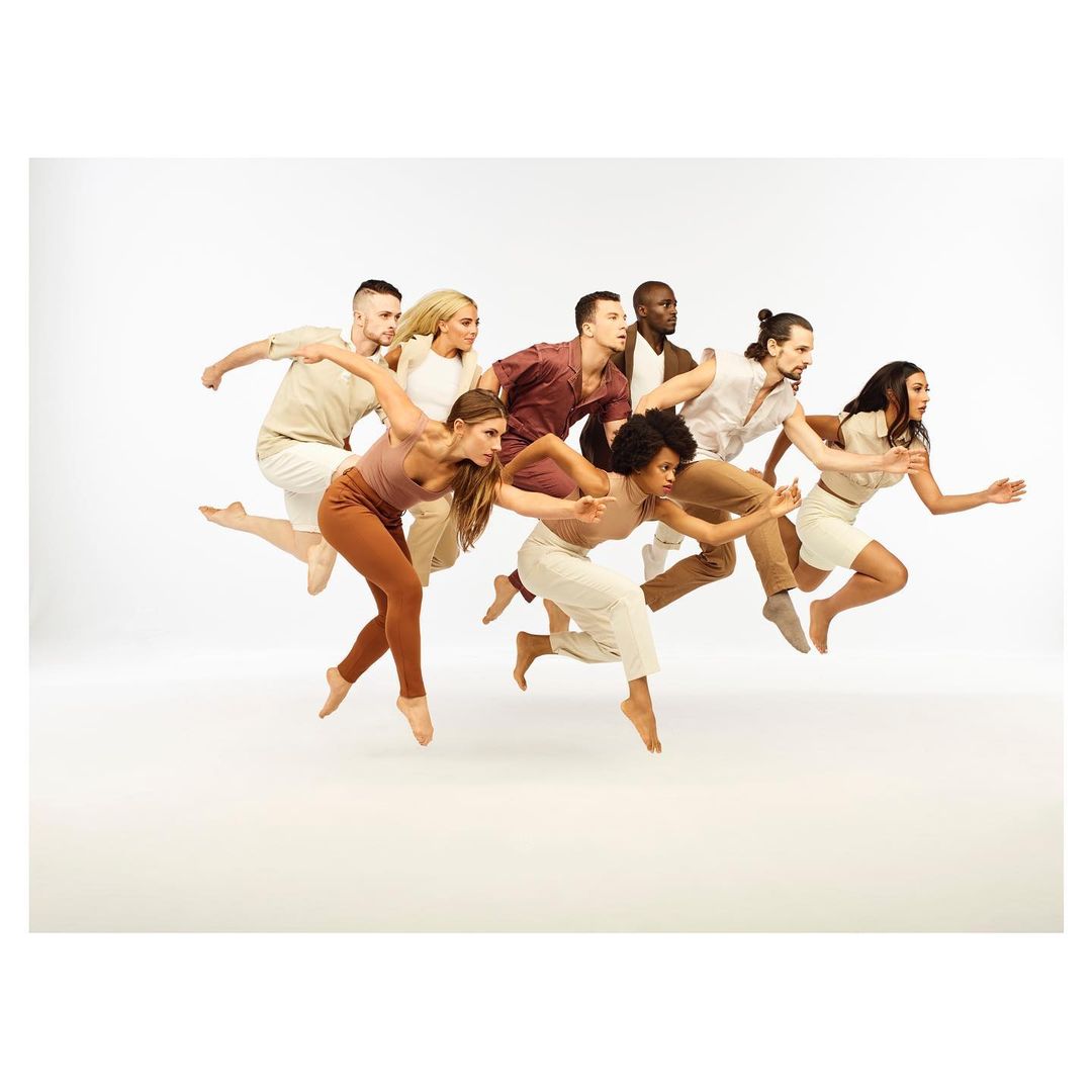 Group of dancers in motion against a white backdrop, wearing earthy-toned clothing. Contemporary dance performance with diverse participants.