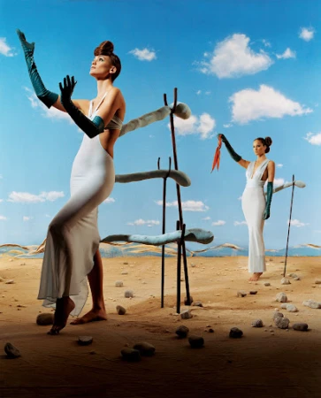 Two women in white dresses and teal gloves pose in a surreal desert landscape with a blue sky and unique sculptural props. Fashion photography.