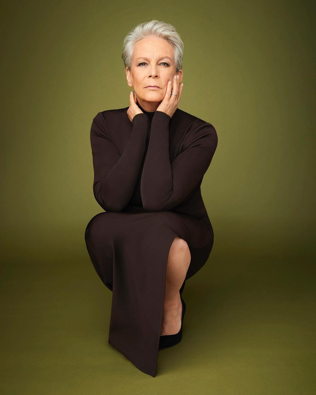 Full-length portrait of Jamie Lee Curtis in a brown dress crouching with hands on her face against a green backdrop, shot in a photo studio.