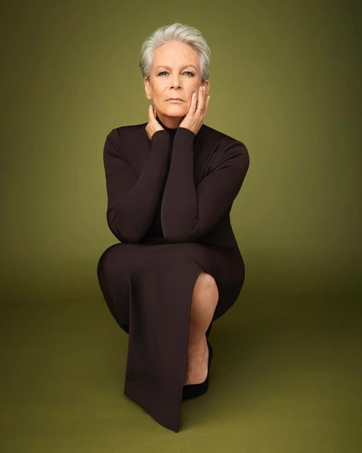 Full-length portrait of Jamie Lee Curtis in a brown dress crouching with hands on her face against a green backdrop, shot in a photo studio.