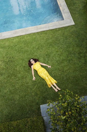 Overhead shot of a woman in a yellow lace dress lying on green grass near a turquoise swimming pool on a sunny day.