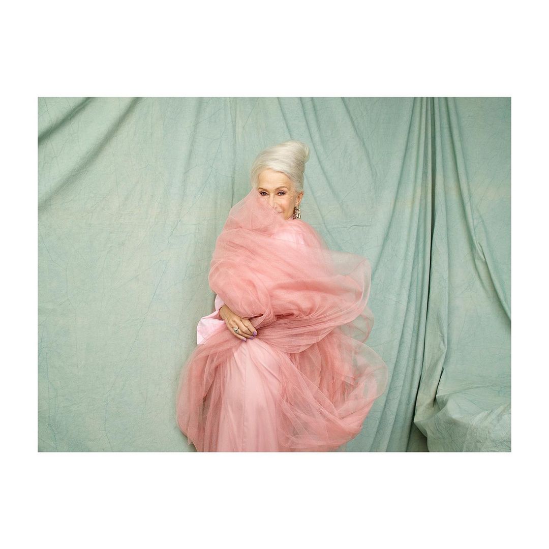 Mature woman in an elegant pink dress posing in front of a teal draped backdrop with tulle fabric partially covering her face.
