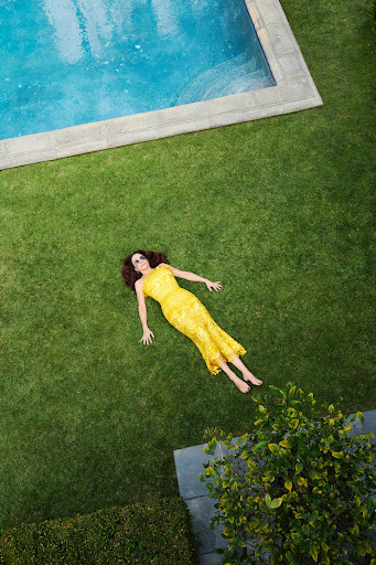 Overhead view of a woman in a yellow dress lying on green grass, near a rectangular pool with blue water and concrete edging.