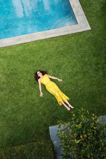 Overhead view of a woman in a yellow dress lying on green grass, near a rectangular pool with blue water and concrete edging.