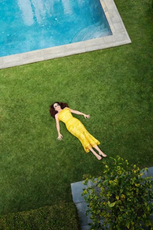 Overhead view of a woman in a yellow dress lying on green grass, near a rectangular pool with blue water and concrete edging.