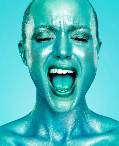 Close-up studio shot of a blue-painted adult woman with closed eyes and mouth open wide, tongue visible, against a light blue background.