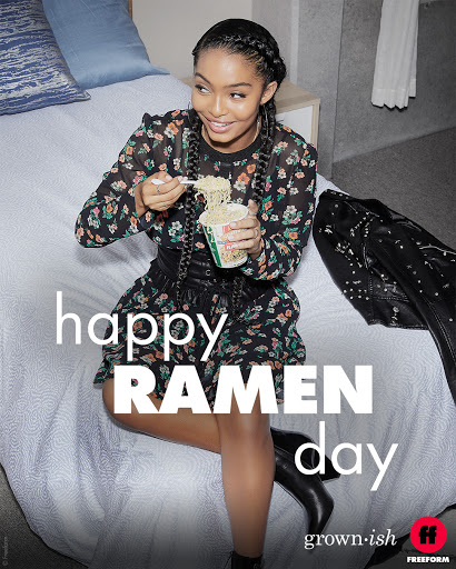 Young woman in floral dress and black boots sits on a bed eating noodles with a fork indoors against a wall and a black jacket.