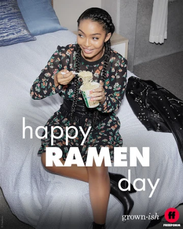 Young woman in floral dress and black boots sits on a bed eating noodles with a fork indoors against a wall and a black jacket.