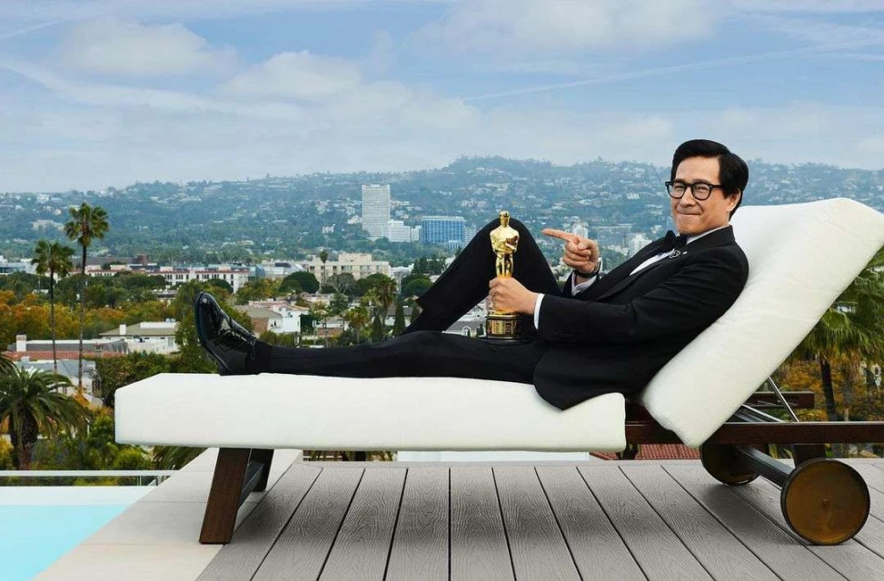 Asian adult male in suit, reclining with an Oscar trophy on a white lounge chair, with a cityscape backdrop on a sunny day.