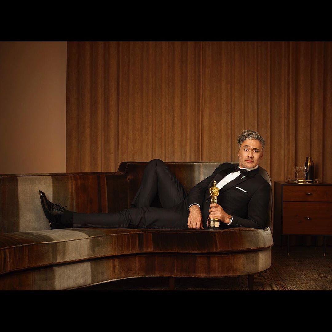 Portrait of a man in a tuxedo reclining on a couch and holding an Oscar award, set against a neutral-toned backdrop.