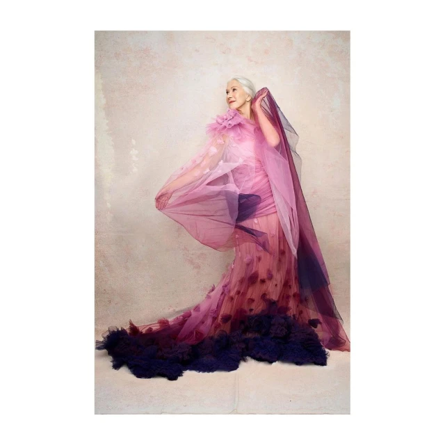Elderly woman poses in a flowing, ombré tulle gown in a photo studio against a textured beige backdrop, creating an elegant portrait.