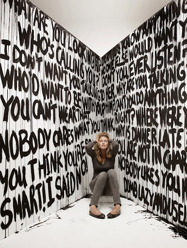 Woman sits in corner surrounded by graffiti-style text. She has her hands on her head with closed eyes, and appears overwhelmed.