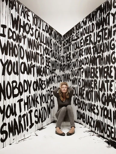 Woman sits in corner surrounded by graffiti-style text. She has her hands on her head with closed eyes, and appears overwhelmed.