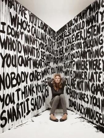 Woman sits in corner surrounded by graffiti-style text. She has her hands on her head with closed eyes, and appears overwhelmed.