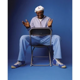 Samuel L. Jackson smiling while seated backward on a folding chair against a solid blue background wearing light blue track jacket and pants