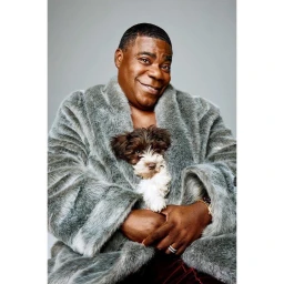 Tracy Morgan wearing a grey faux fur robe holding a small brown and white puppy against a light grey studio background.