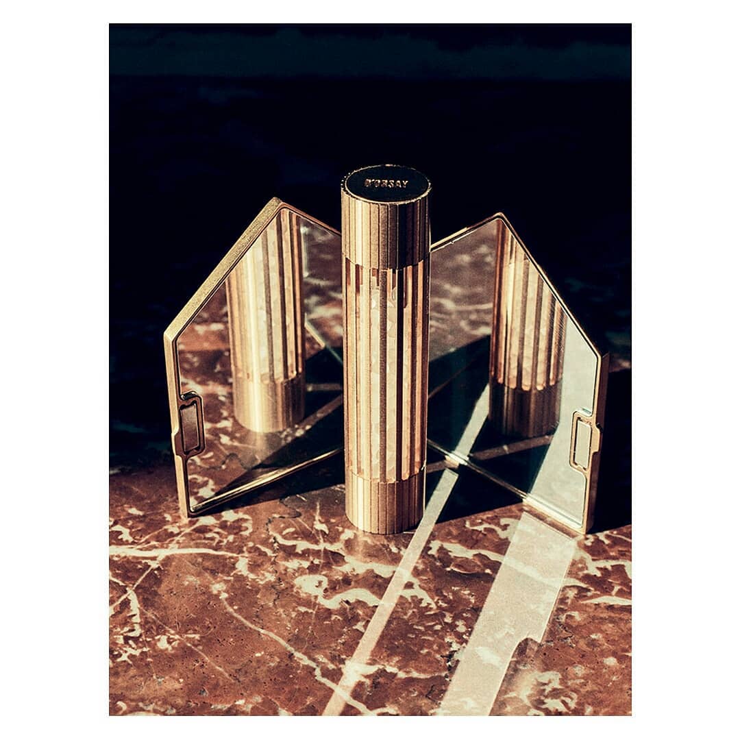 Still life shot of a gold cylindrical perfume bottle with textured vertical ridges, reflected in mirror on a marble surface.