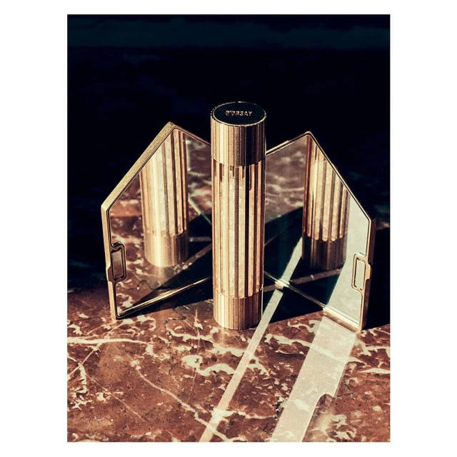 Still life shot of a gold cylindrical perfume bottle with textured vertical ridges, reflected in mirror on a marble surface.