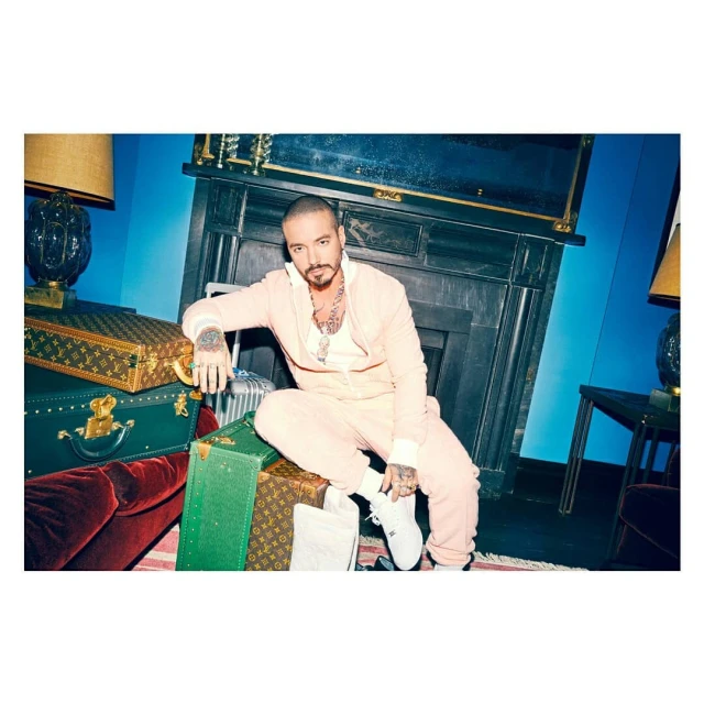 J Balvin, in a peach tracksuit and white sneakers, poses on vintage luggage in an indoor studio setting with a blue wall and fireplace.
