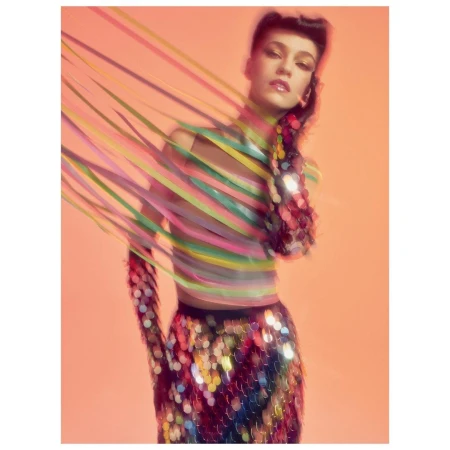 Woman in sequin skirt and colorful ribbon wraps, posing in a studio with coral background. Fashion portrait in soft focus.