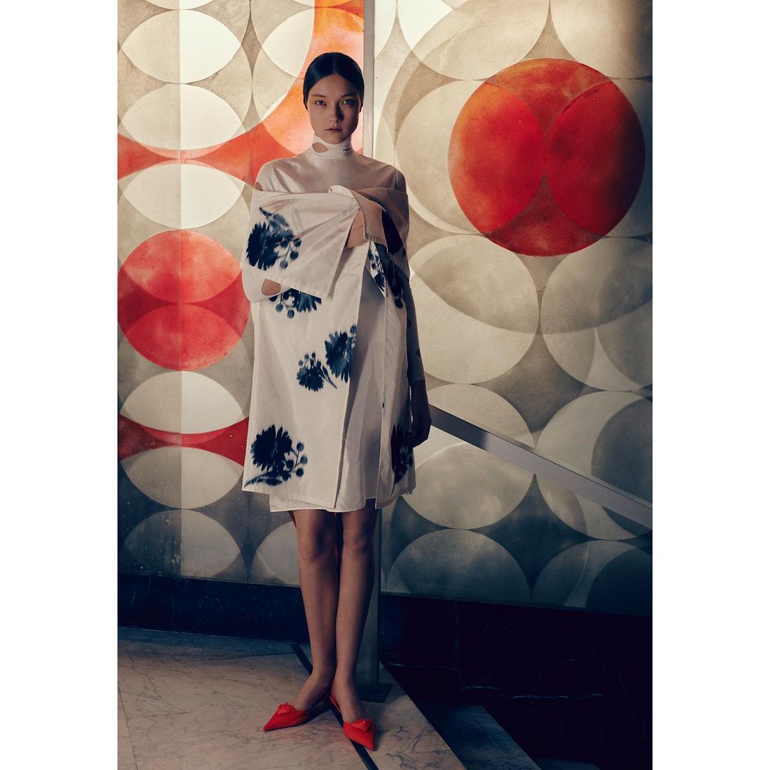 A stylish woman poses in a white floral dress and red shoes in front of a geometric patterned wall, fashion-forward and elegant.