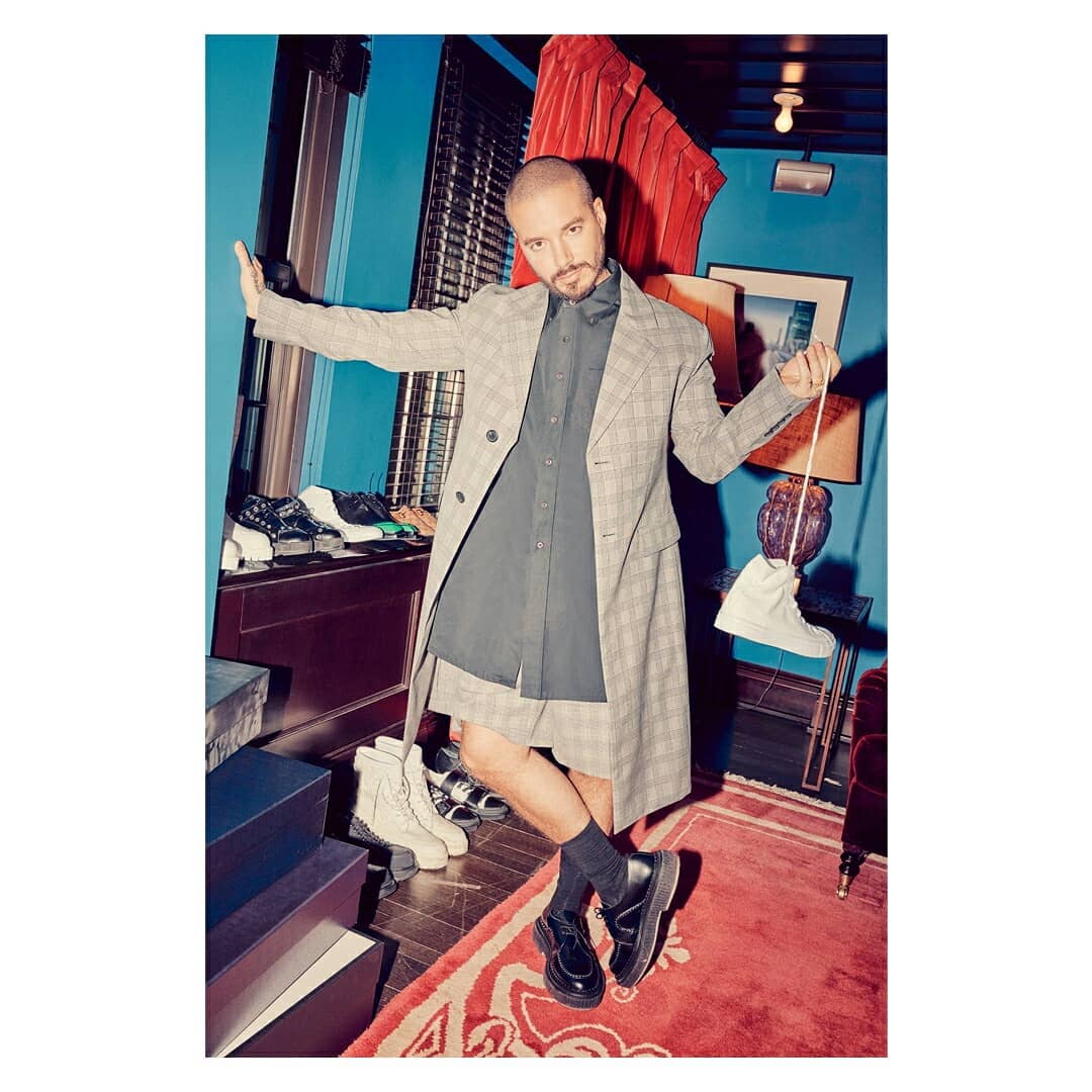 Stylish man in gray plaid coat, black shirt, and shorts posing with shoes in an eclectic room featuring turquoise walls and red rug.