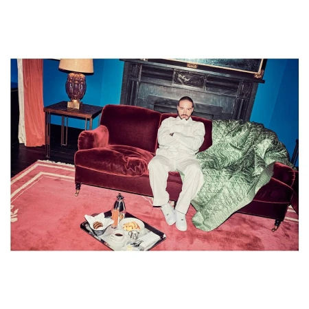 Adult male seated on a burgundy couch in a room with breakfast tray and a fireplace, wearing an off-white jumpsuit and sneakers.