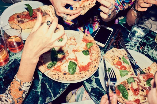 Overhead shot of people sharing pizza and appetizer on a marble table, glasses of drinks, a phone, and silverware present.