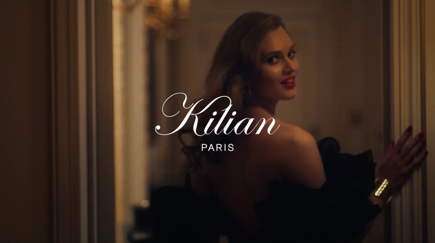 A woman with red lipstick, wearing a black dress and gold bracelet, poses near a doorway with 'Kilian Paris' logo in white.