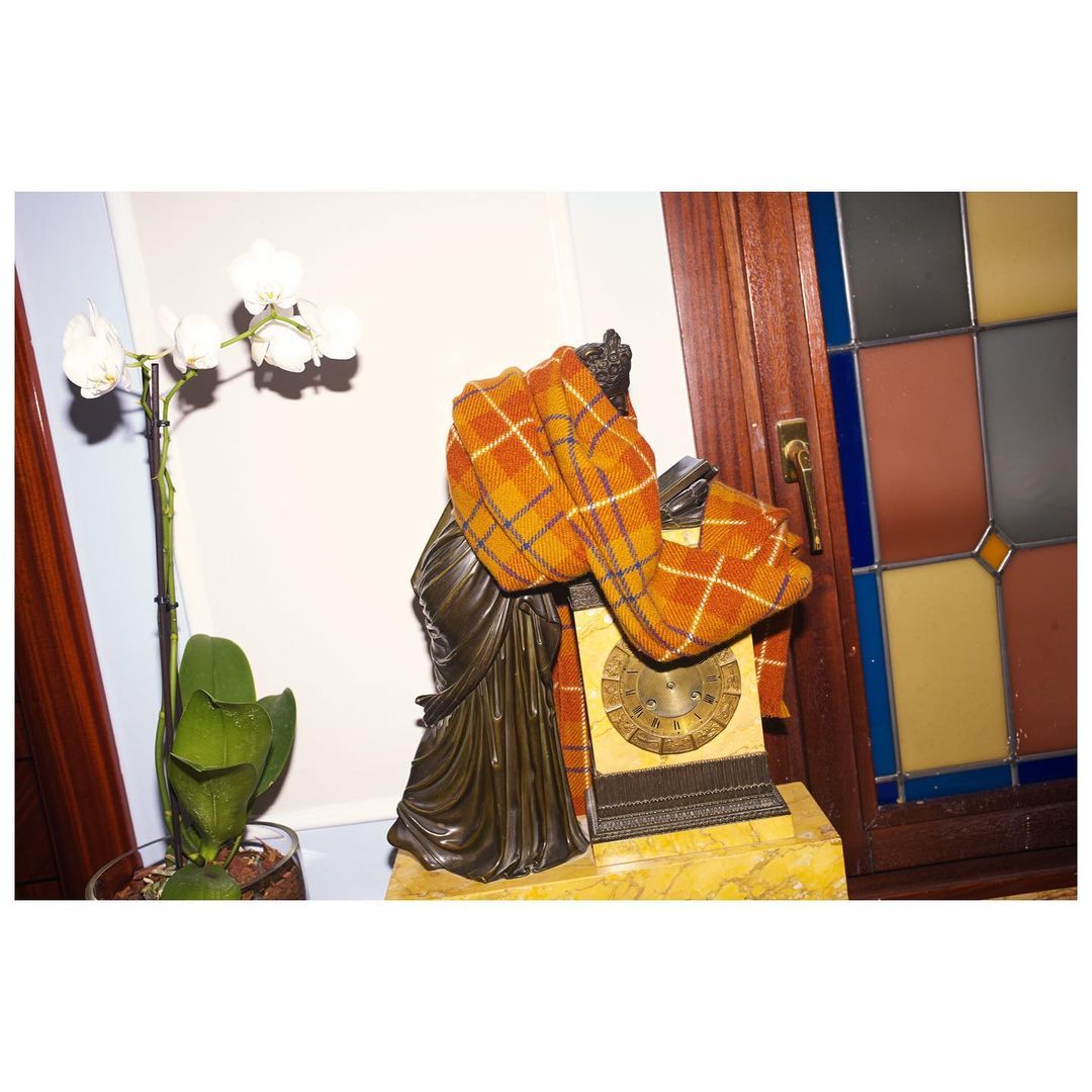 Still life featuring a bronze statue draped with a plaid scarf, an antique clock, and an orchid plant in a softly lit indoor setting.