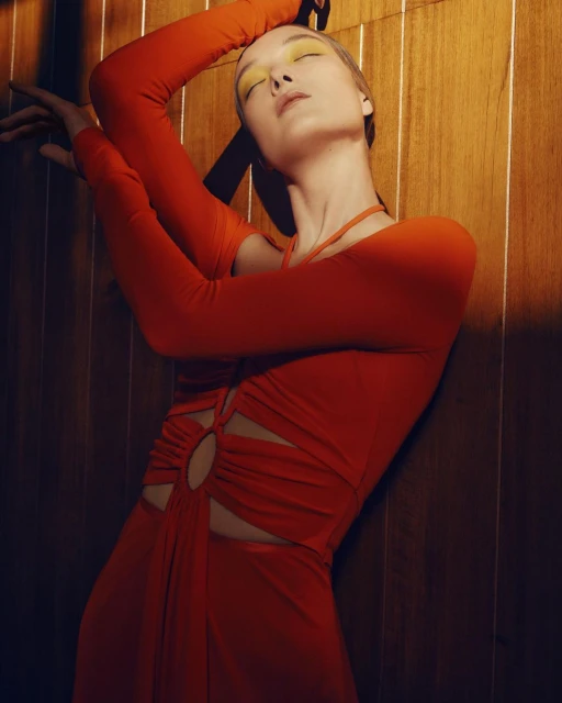 Fashion shot of a woman in a red dress leaning against a wood wall, eyes closed, yellow eyeshadow, long sleeves, studio lighting.