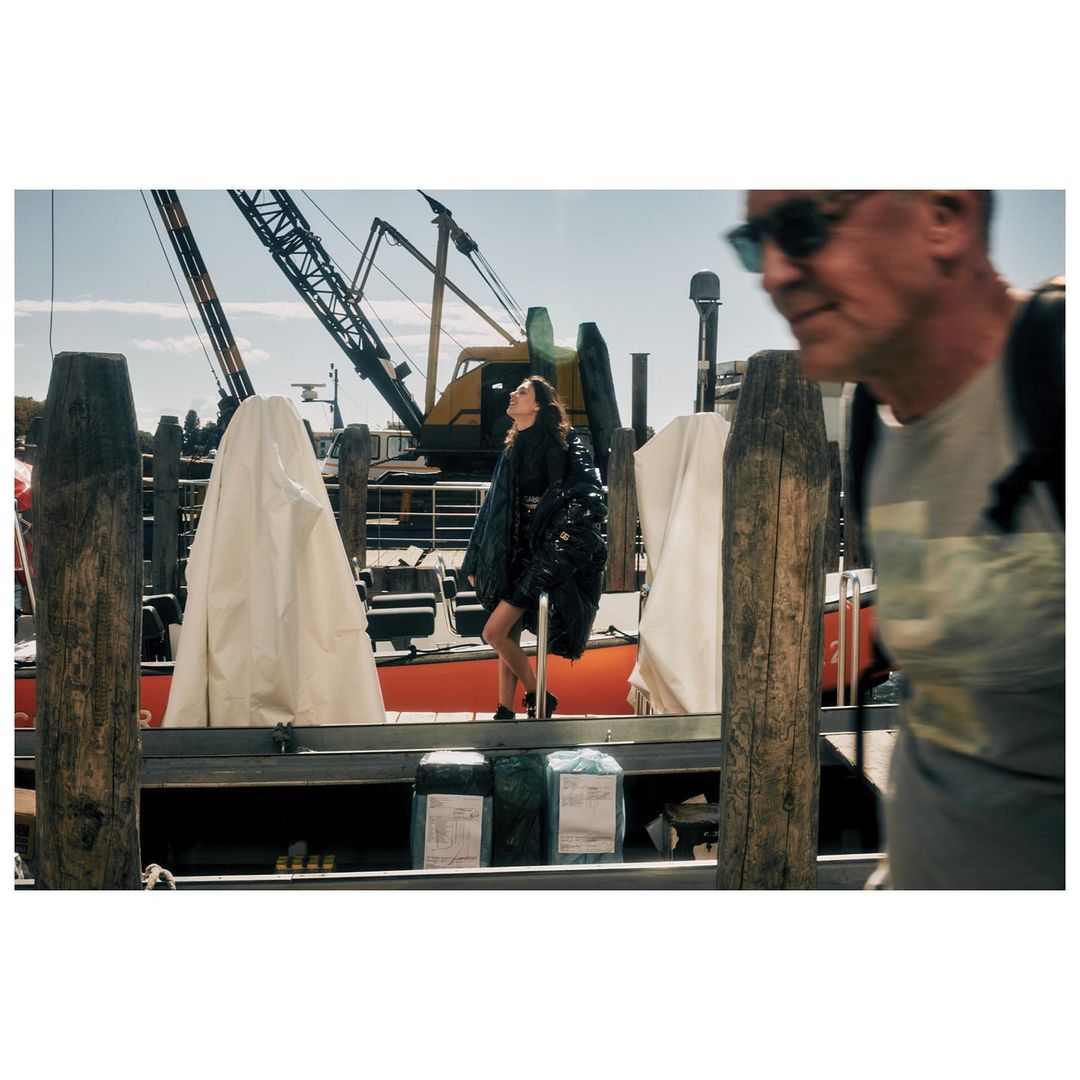 Woman in a shiny jacket stands on a pier near a crane and other people on a sunny day. Fashion editorial style photography.