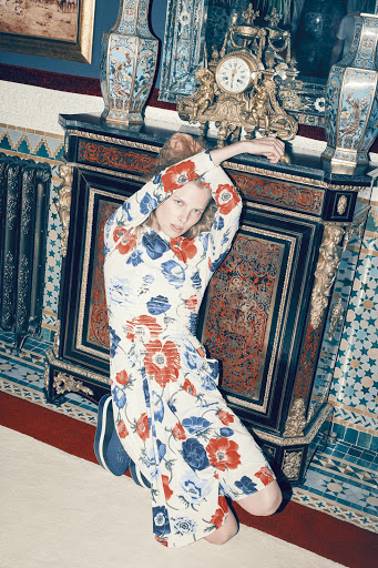 A woman in a floral print dress poses in front of an ornate dark wood and gilt cabinet with a clock and vases.